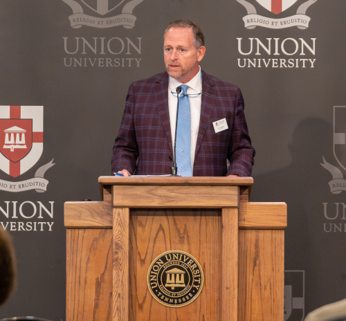 A man in a checked blazer and blue tie stands at a wooden podium with the Union University seal, addressing gospel outreach efforts in front of a backdrop displaying the Union University logo.
