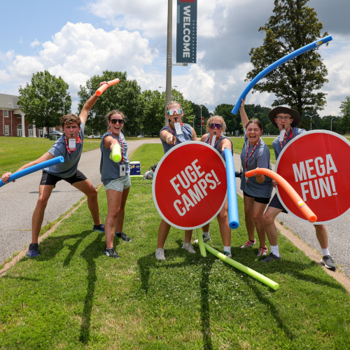 Six people stand outside on grass holding pool noodles and large signs reading FUGE CAMPS! and MEGA FUN! The group, serving as friendly Union hosts, poses energetically in front of a WELCOME banner.
