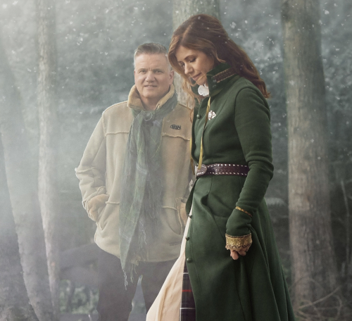 A woman in a long green coat stands in the foreground looking down, while a man in a tan jacket stands behind her, both surrounded by fog in a wooded area—capturing the quiet mood before the Union Scholarship Banquet.