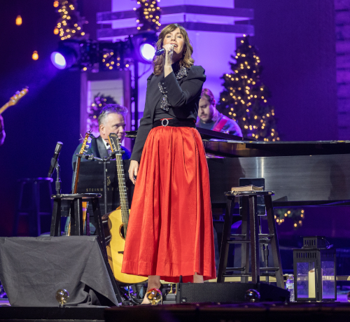 A woman in a black top and long red skirt sings into a microphone on stage at the Union Scholarship Banquet, with holiday lights, decorated trees, and the Getty duo playing instruments nearby.