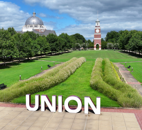 Large white letters spelling UNION sit on a paved walkway in front of a grassy area, clock tower, and domed building—where Union grad students gather beneath rows of trees under a partly cloudy sky.