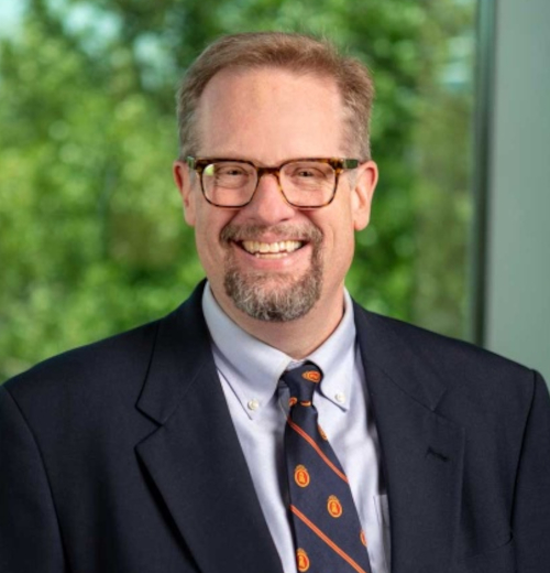 A man with short light brown hair, glasses, and a beard smiles while wearing a dark suit, white shirt, and patterned tie. He stands indoors in front of a window with greenery visible outside at the May 4 commencement for Union graduates.