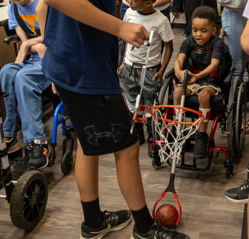 A group of children, some in wheelchairs, gather indoors as a boy holds a toy basketball hoop close to a basketball. Inspired by the Make:able challenge, one child in a wheelchair appears ready to play on the wooden floor with local student teams nearby.