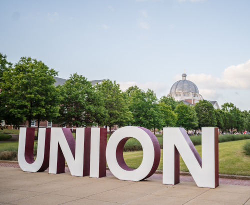 Large block letters spell UNION in front of green trees and a domed building under a blue sky with scattered clouds on this Christian college campus, recognized as one of the best colleges by Niche.