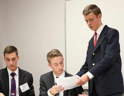 Three young men in suits are in a meeting room during a debate tournament. One stands and hands a paper to a seated colleague, while the third looks down at the table. A whiteboard is visible in the background.