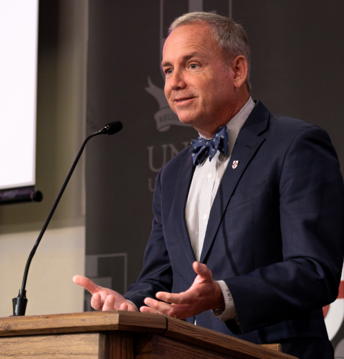 A man in a navy suit, bow tie, and striped shirt—possibly Oliver—speaks at a podium with a microphone, gesturing as he discusses godly leadership before West Tennessee pastors against a dark backdrop with faint text and logos.