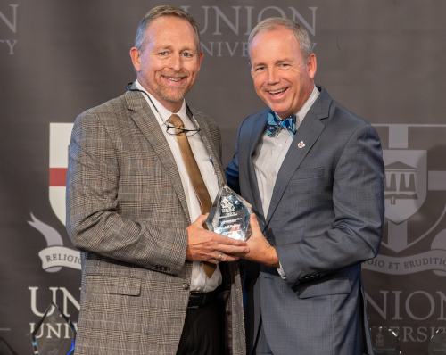 Two men in suits stand side by side, smiling at the camera. One is handing the other a triangular glass award for Alumnus of the Year during Union Homecoming, with a backdrop displaying the Union University and Perkins logos.