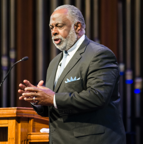 An older man with gray hair and a beard, identified as Smith, speaks at a podium during the Dockery Lectures. He is wearing a dark pinstripe suit, light blue tie, and pocket square. Organ pipes are visible in the background on March 25.
