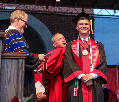 A graduate in cap and gown smiles while receiving an academic hood from a faculty member at Union spring commencement. Another faculty member stands nearby, holding a program and smiling, as students receive degrees.