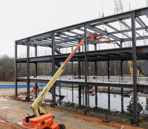A construction site with a multi-story steel frame structure being built. Workers in union safety gear operate lifts and cranes, with building materials and reflective wet ground visible. Trees are in the background.