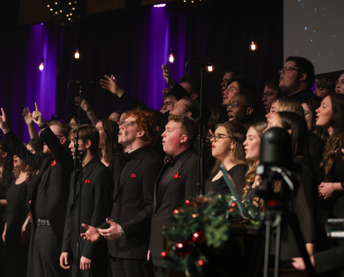 A choir dressed in black formal attire sings on stage at A Union Christmas concert, with some members raising their hands. Microphones and festive decorations are visible, and string lights hang in the background for the Dec. 6 event.