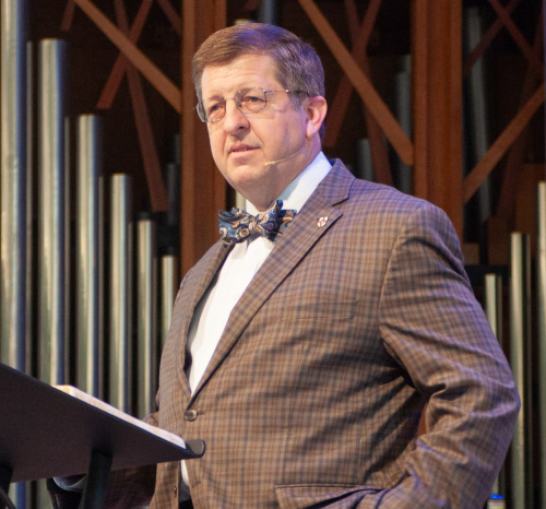 A man wearing glasses, a plaid suit, and a bow tie stands at a podium, speaking. Behind him are large vertical metal pipes, suggesting the setting is a church or auditorium. He is likely Van Neste, vice president for university ministries at Union.