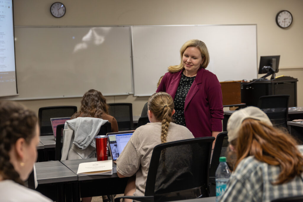 A woman in a maroon blazer stands and talks to students seated at desks with laptops in a classroom. There are clocks, a whiteboard, and a projector screen visible in the background.