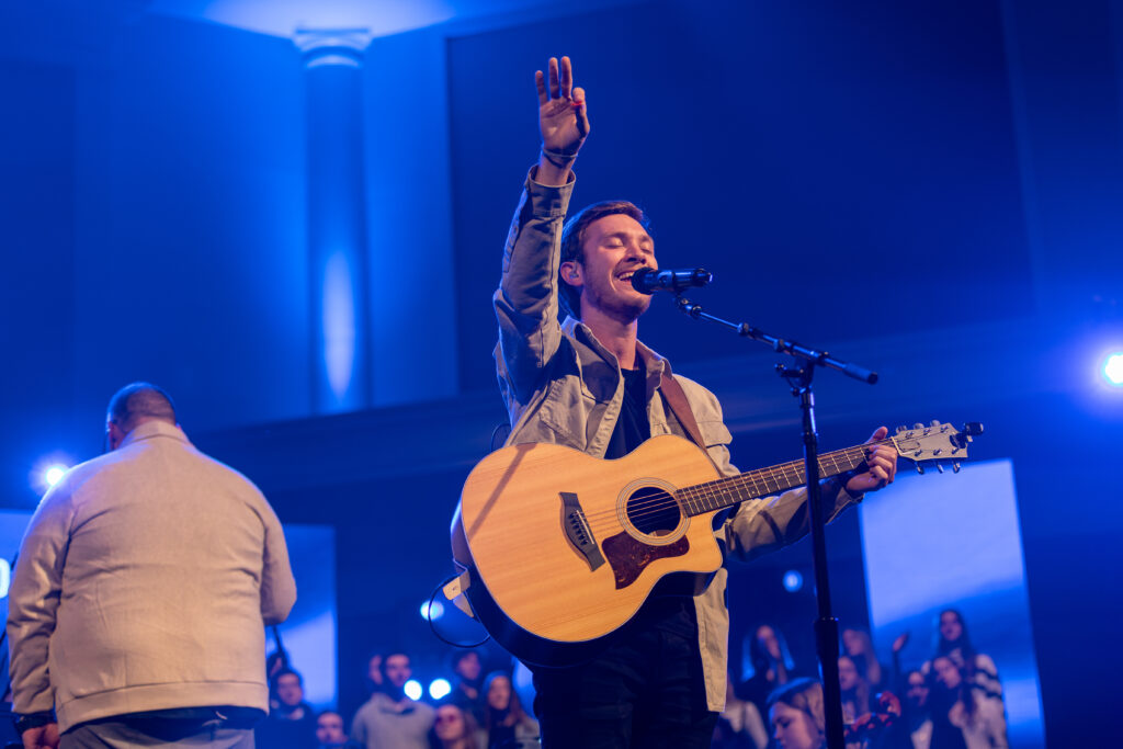 A musician from the Department of Music stands on stage playing an acoustic guitar and singing into a microphone with one arm raised. Blue lighting fills the venue, and a crowd is visible in the background.