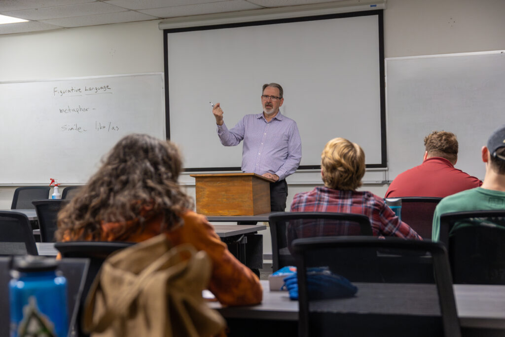 A man stands at a lectern teaching a classroom of seated students. Notes on writing and literature are written on the whiteboard behind him. The students face forward, listening to the lesson attentively.