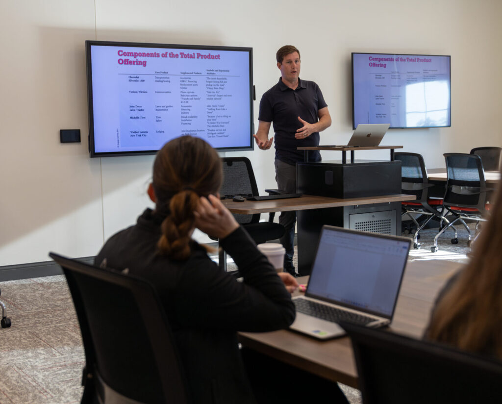 A person stands at the front of a conference room giving a presentation on Components of the Total Product Offering, with slides displayed on two screens. Attendees sit at desks with laptops and take notes.