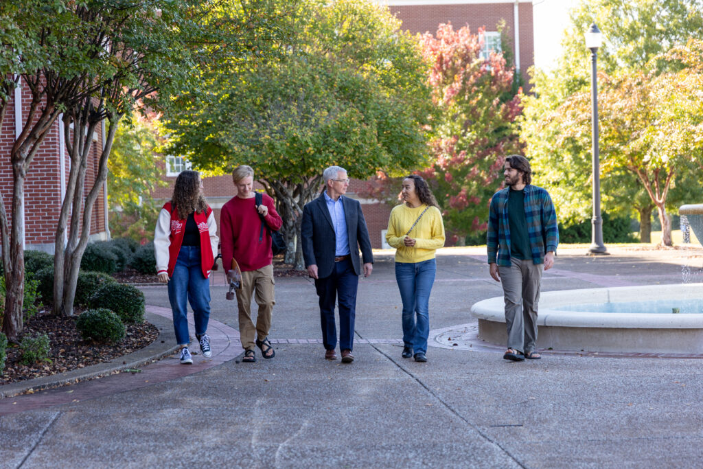 Five people walk together on a campus pathway lined with trees and a fountain, likely discussing topics from the Department of History. They appear engaged in conversation, with autumn foliage visible in the background.