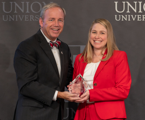 Two people stand in front of a Union University backdrop during annual Homecoming week. The man in a dark suit and bow tie presents the Alumnus of the Year award to the woman in red, both smiling at the camera.