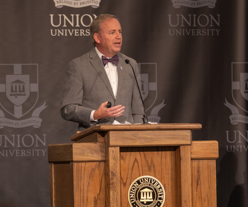 A man in a gray plaid jacket and purple bow tie stands at a wooden podium with the Union University logo, delivering an institutional statement on biblical anthropology to union trustees, before a backdrop displaying the Union University name and crest.