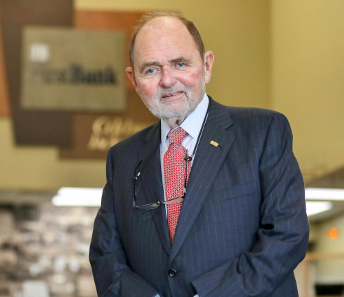 An older man with a beard and short hair, known as a campus champion, wears a dark pinstripe suit, red tie, and glasses hanging from his collar as he stands indoors in front of blurred union wall signage.