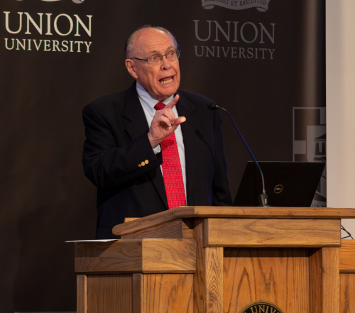 An older man in a suit and red tie speaks at a wooden podium with a laptop on it during a Bible conference. The backdrop features the words Union University and University logos.