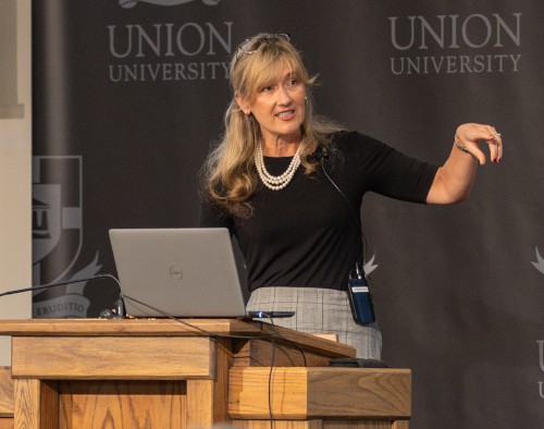A woman stands at a wooden podium with a laptop, wearing a black top, gray skirt, and pearl necklace, gesturing with her right hand. The background shows Union University banners, highlighting the Union community’s interest in Civil War history.
