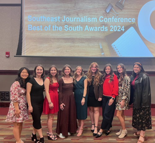 Nine women, all C&C staffers, stand in a row, posing and smiling for a group photo at the Southeast Journalism Conference Best of the South Awards 2024, with a presentation slide behind them highlighting the SEJC competition.