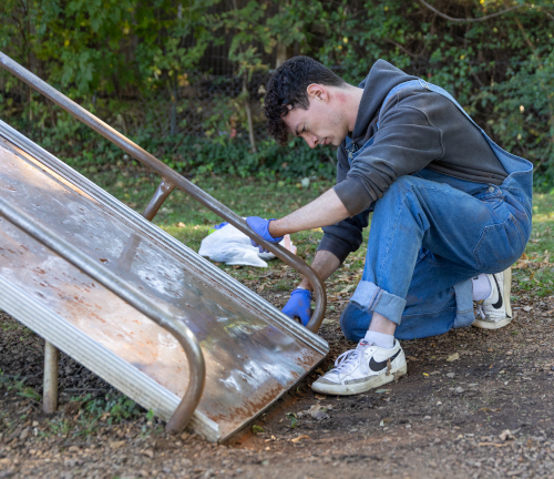 A local resident wearing blue gloves and jeans crouches next to a metal slide outdoors, cleaning or inspecting its surface. Fallen leaves cover the ground, with greenery in the background—capturing a community spirit on Campus and Community Day.