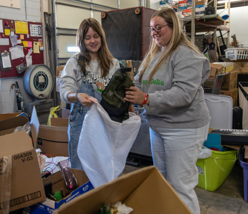 Two women stand in a warehouse at the Jackson Union, sorting items into a large white bag. Surrounded by open boxes, they appear to be organizing belongings, likely preparing for Campus and Community Day.