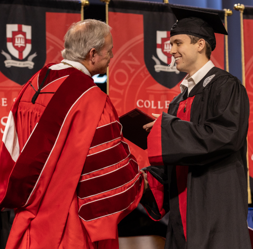 A Union graduate in a black cap and gown shakes hands with a university official in red academic regalia while receiving a diploma on stage, celebrating the completion of a hard path. Red banners with a school emblem hang in the background.