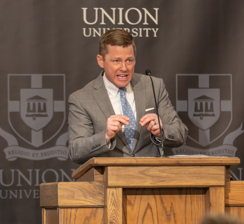 A man in a gray suit and blue polka-dot tie speaks emphatically at a wooden podium during the Dockery Lectures. The backdrop displays the Union University logo and motto, Religio et Eruditio.