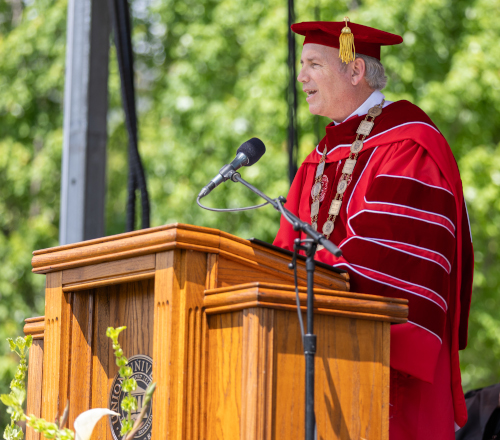 Oliver, dressed in academic regalia with a red robe and cap, stands at a wooden podium speaking into a microphone during graduation. The outdoor ceremony is set against a backdrop of green trees on Union grounds.