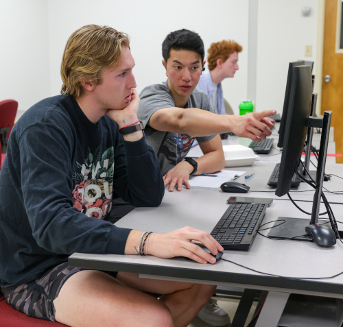 Three students sit at desks with computers in a classroom. One points at a monitor while another, studying computer engineering, uses a mouse. In the background, a third student works quietly, focused on their screen.