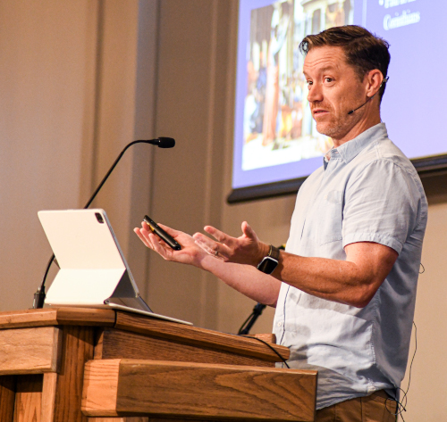 A man stands at a podium with a laptop and microphone, gesturing as he addresses the EQUIP Youth Apologetics Conference. He holds a phone in one hand while speaking in front of a projected presentation slide at Union.