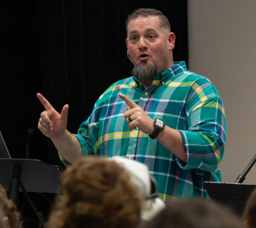 A man with a goatee wearing a colorful plaid shirt gestures with both hands while speaking to an audience at the 4th annual Youth Apologetics Conference in an indoor setting.