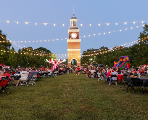 People sit at tables on a lawn during Family Weekend at Union, enjoying an outdoor event at dusk. String lights glow overhead, a tall clock tower rises in the background, and a bounce house and carnival rides set the scene for Sept. 26-27.