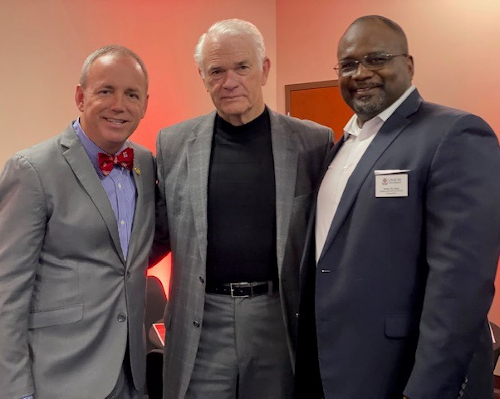 Three men in suits stand together indoors at the UU Germantown anniversary, posing for a photo. The man on the left sports a bow tie, the middle wears a turtleneck under a blazer, and the right has glasses and a name badge.