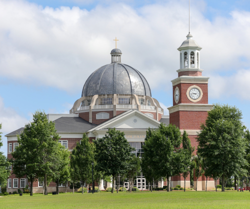 A large brick building with a prominent domed roof topped by a cross stands next to a tall clock tower, where volunteers gather for the Union's Scripture reading marathon amid green trees and grass under a partly cloudy sky.