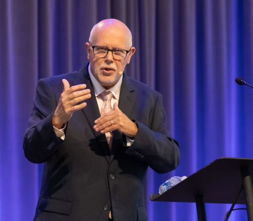 A bald man with glasses, identified as retired professor Moore, is wearing a suit and tie as he speaks and gestures behind a podium on stage, with purple curtains in the background at the Union graduation.