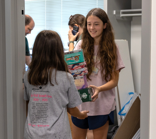 Two girls stand in a doorway on Move-In Day 2025; one hands a stack of boxes to the other. Both wear casual clothes. Two adults are visible in the background, one holding a camera, near a closet filled with various items.