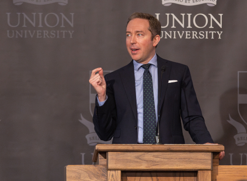 A man in a suit and tie speaks at a podium in front of a backdrop with the words Union University and a university crest, gesturing with his right hand as he discusses work within the context of God’s story.