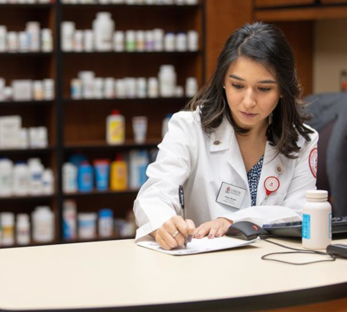A woman in a white lab coat writes on a clipboard at a counter in a pharmacy, where Union pharmacy students in Tennessee prepare for high NAPLEX pass rates, with medicine bottles and shelves of medications visible in the background.