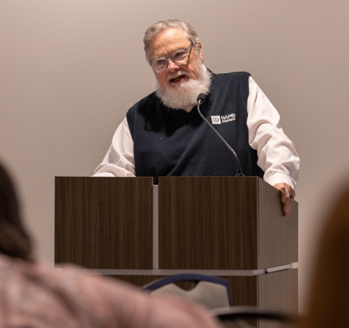 An older man with a white beard and glasses stands behind a wooden podium, speaking. He is wearing a white shirt and a dark NAMM vest. The background is plain, and some local pastors are visible in the foreground.