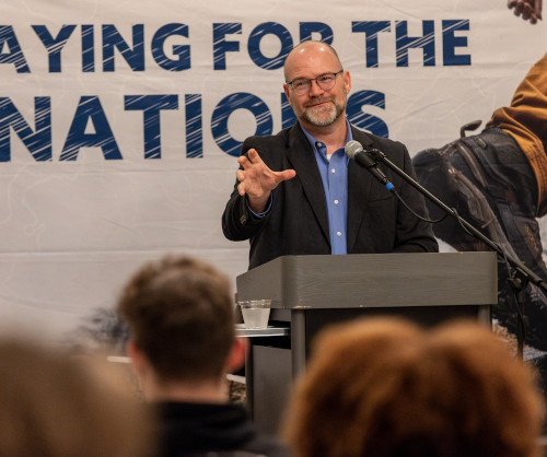 A man in a suit and glasses stands at a podium speaking into a microphone, gesturing with his hand. An audience listens as he discusses missions success. A banner behind reads PRAYING FOR THE NATIONS.