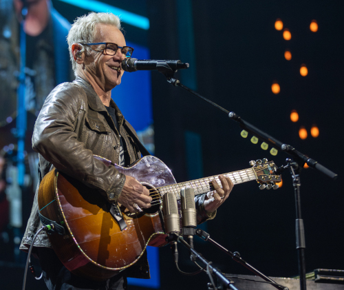 A man wearing glasses and a brown leather jacket smiles while playing an acoustic guitar and singing into a microphone on stage at the Chapman Scholarship Banquet. Stage lights and equipment are visible in the background.