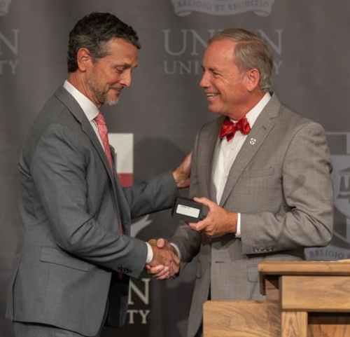 Two men in suits shake hands on a stage as one hands a small box to the other, both smiling. A podium and a Union University backdrop are visible, celebrating the Lady Bulldogs’ Final Four achievement in front of Trustees.