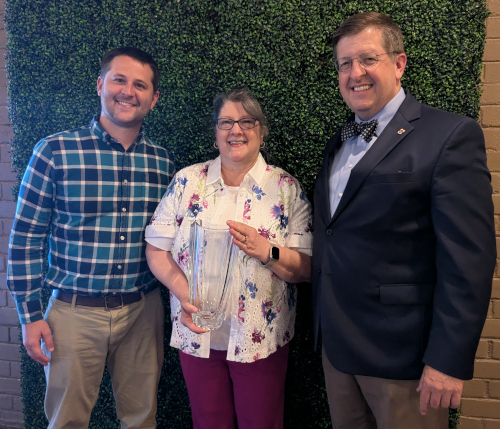 Three people stand together in front of a green leafy wall. The woman in the center holds the clear Dodd Award. The two men on either side are smiling—Shaddix, on the right, wears a suit and bow tie at the SBC meeting.