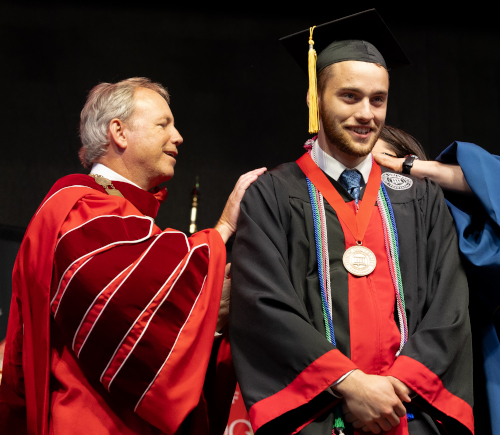 A man in a red academic gown touches the shoulder of a young man in a graduation cap, gown, and red stole, smiling and wearing several honor cords and a medal during Union graduation 2024.