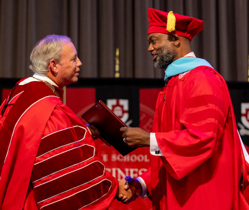 Two men in red academic regalia shake hands on stage at a Union summer graduates ceremony. One holds a diploma cover, both smiling, with a red and black banner in the background as they proudly hold the line of tradition.