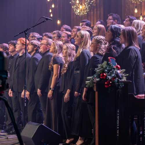 A choir of men and women stands on stage, dressed in black, singing under hanging lights at A Union Christmas concert. A decorated podium with greenery and red ornaments is visible in the foreground, capturing the festive Dec. 5 celebration.